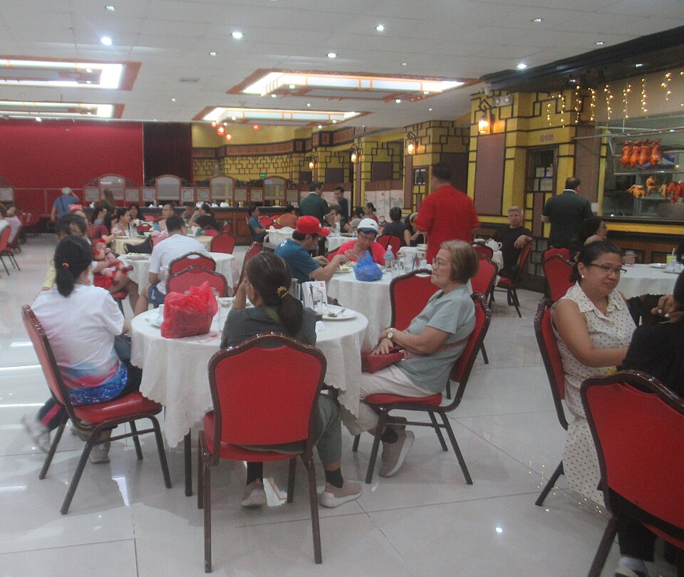Chinese restaurant dining hall with round white-tablecloth tables, red chairs, and diners sharing meals