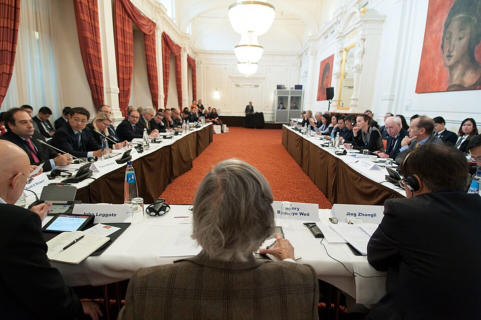 International business conference with Chinese and Western delegates seated around a U-shaped table in an ornate European hall