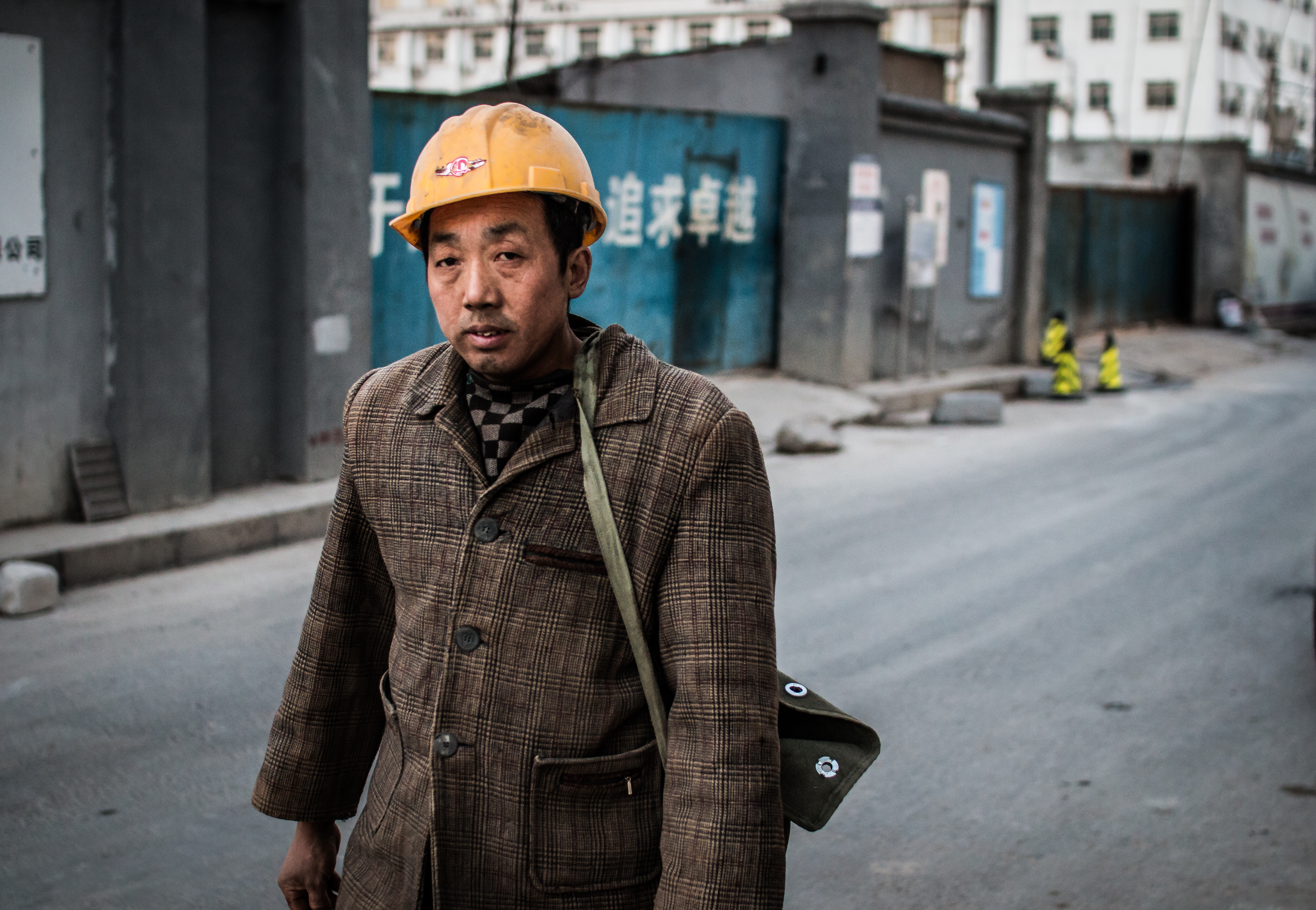 Chinese migrant construction worker wearing a yellow hard hat and carrying tools on a city street with Chinese signage